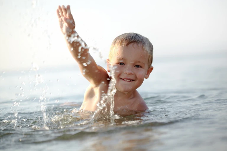  — © Smiling little boy ducking down in the shallow water at the seaside with a gesture of greeting in a tranquil ocean on a summer day