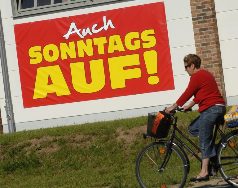 Bäckerei, Verkauf am Sonntag, — © Eine Radfahrerin fährt an einem großen Plakat mit der Aufschrift "Auch Sonntags auf" vorbei. Foto: Stefan Sauer/Archiv